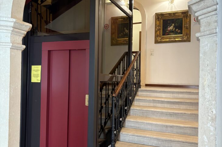 Scala in pietra e ascensore rosso in struttura storica con quadri dorati e pavimento a scacchi