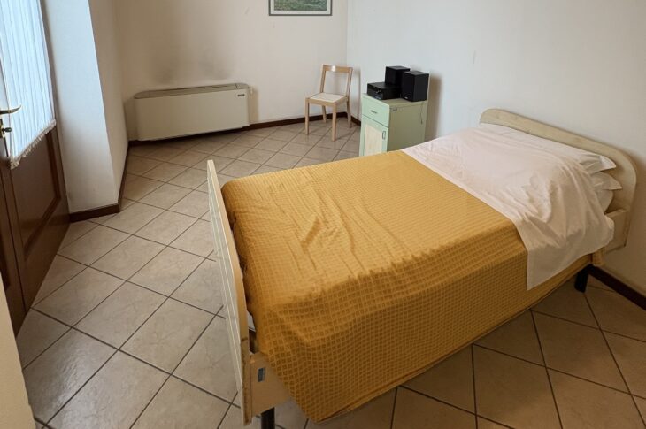 Letto con coperta gialla, stereo, sedia e grande finestra con tapparella verticale