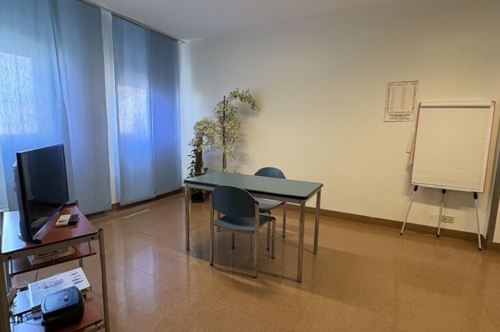 Tavolo con due sedie blu, pianta, lavagna e scaffale con TV e proiettore