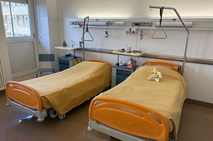 Due letti con coperte beige, uno con peluche, e attrezzature mediche al muro
