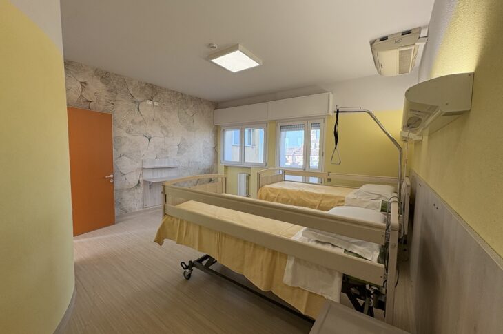 Due letti con sponde e trapezio, pareti chiare e porta arancione con vista esterna