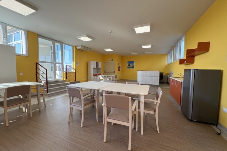 Tavoli e sedie in ambiente giallo con angolo cucina e scala verso il corridoio